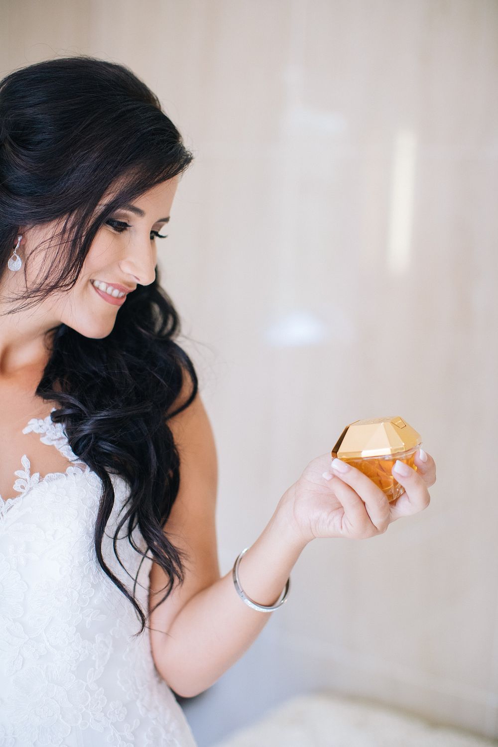 Smiling bride in white lace gown holding a gold diamond-shaped perfume bottle on her wedding day.