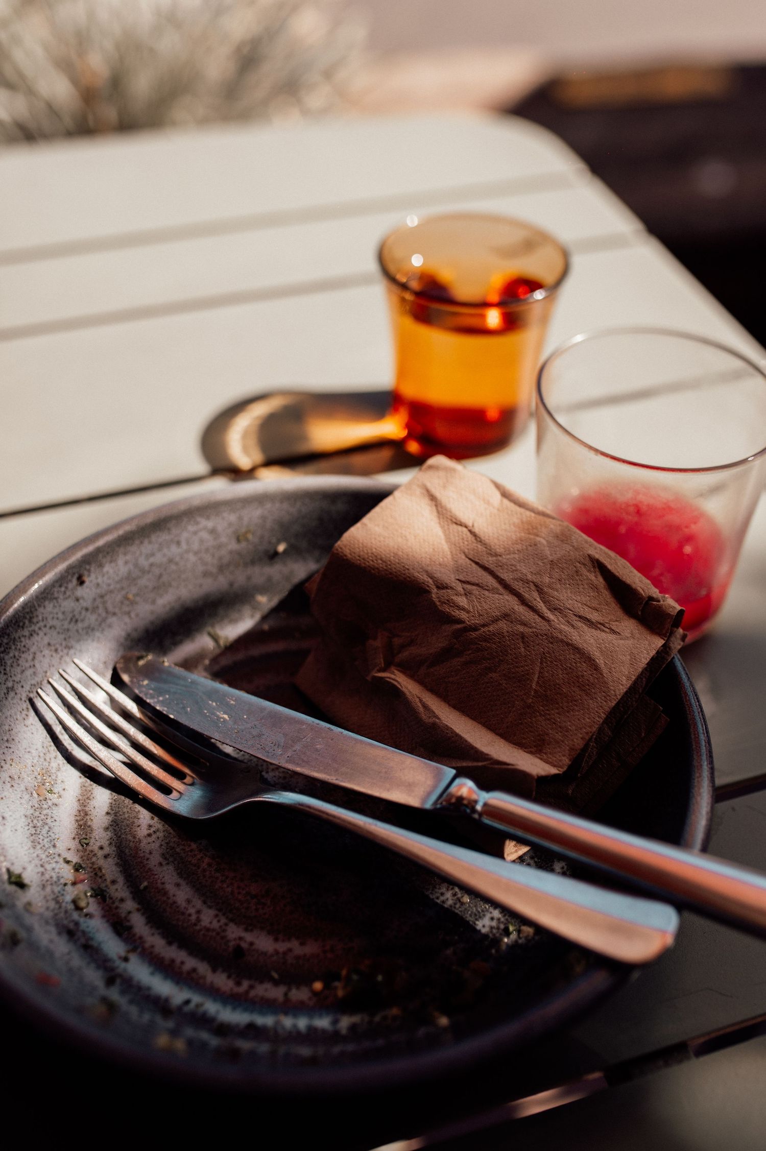 Dark ceramic plate with fork, knife, and brown paper bag on white table with amber glass and red drink.
