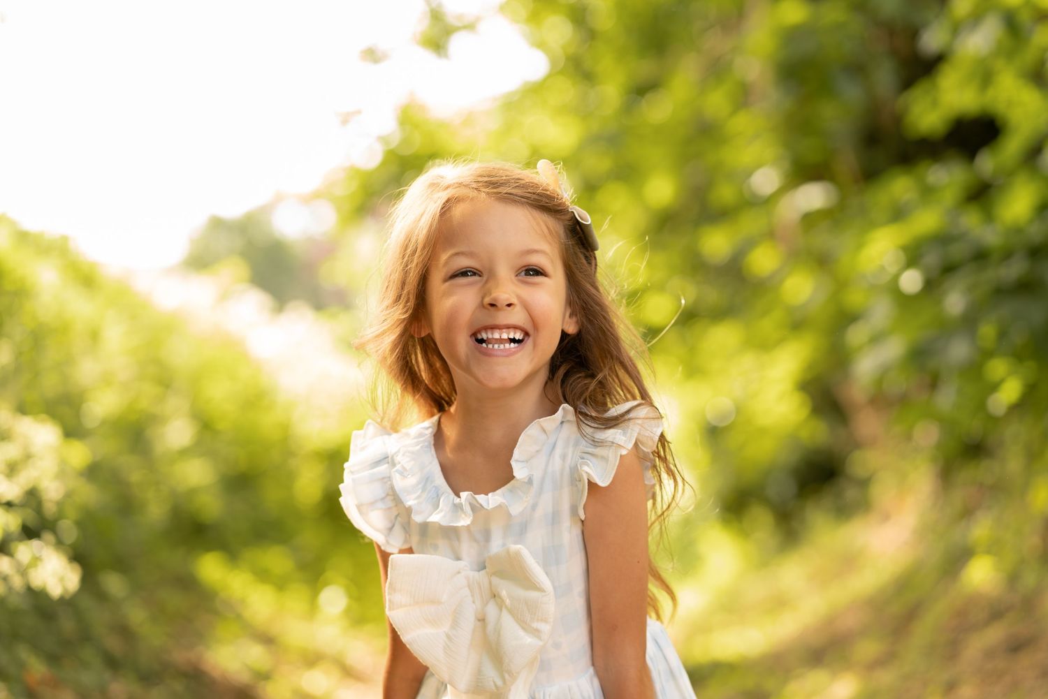 A joyful young girl in a white dress laughs outdoors on a sunny path surrounded by lush green trees.