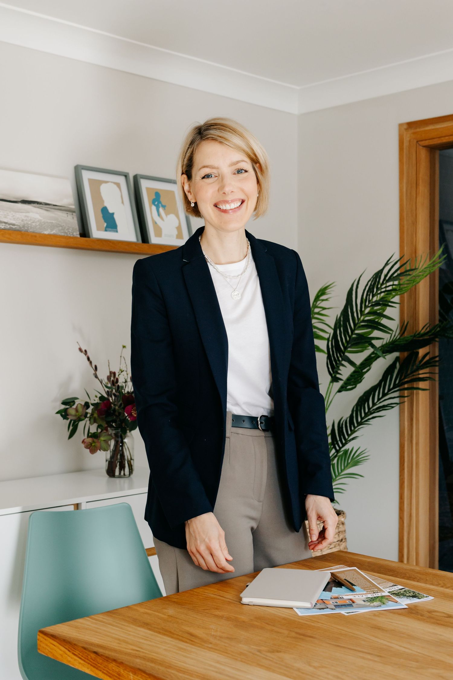 A smiling professional woman in a black blazer stands at a wooden desk in a modern office with framed art and plants.