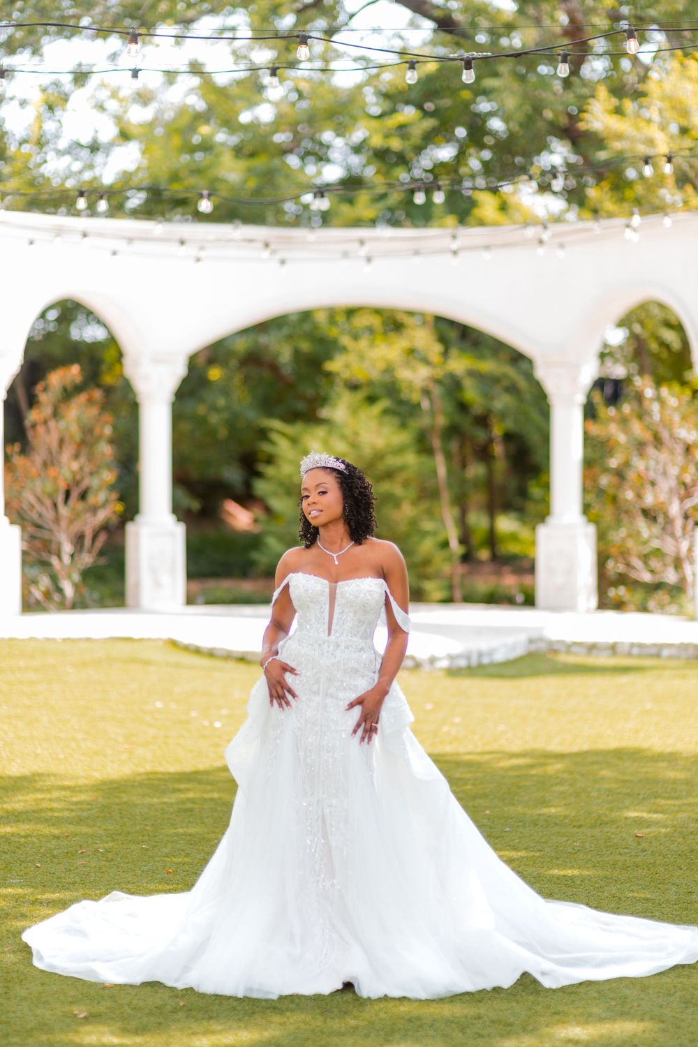Bride in white wedding gown poses gracefully on lawn with elegant white pergola in background during outdoor photo session.