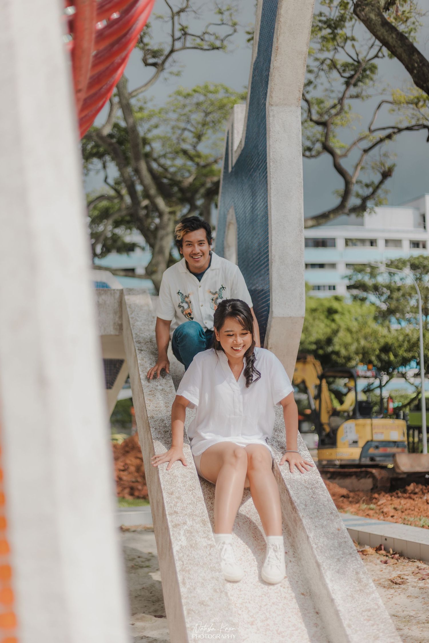 Couple in vintage attire dancing playfully by white gazebo in garden setting.