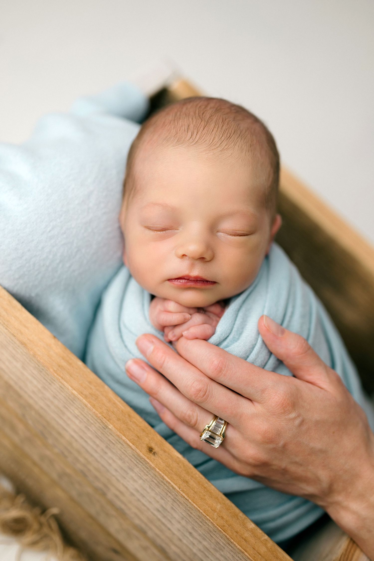 Newborn baby swaddled in light blue wrap sleeping peacefully in wooden crate with adult hand gently resting on chest.