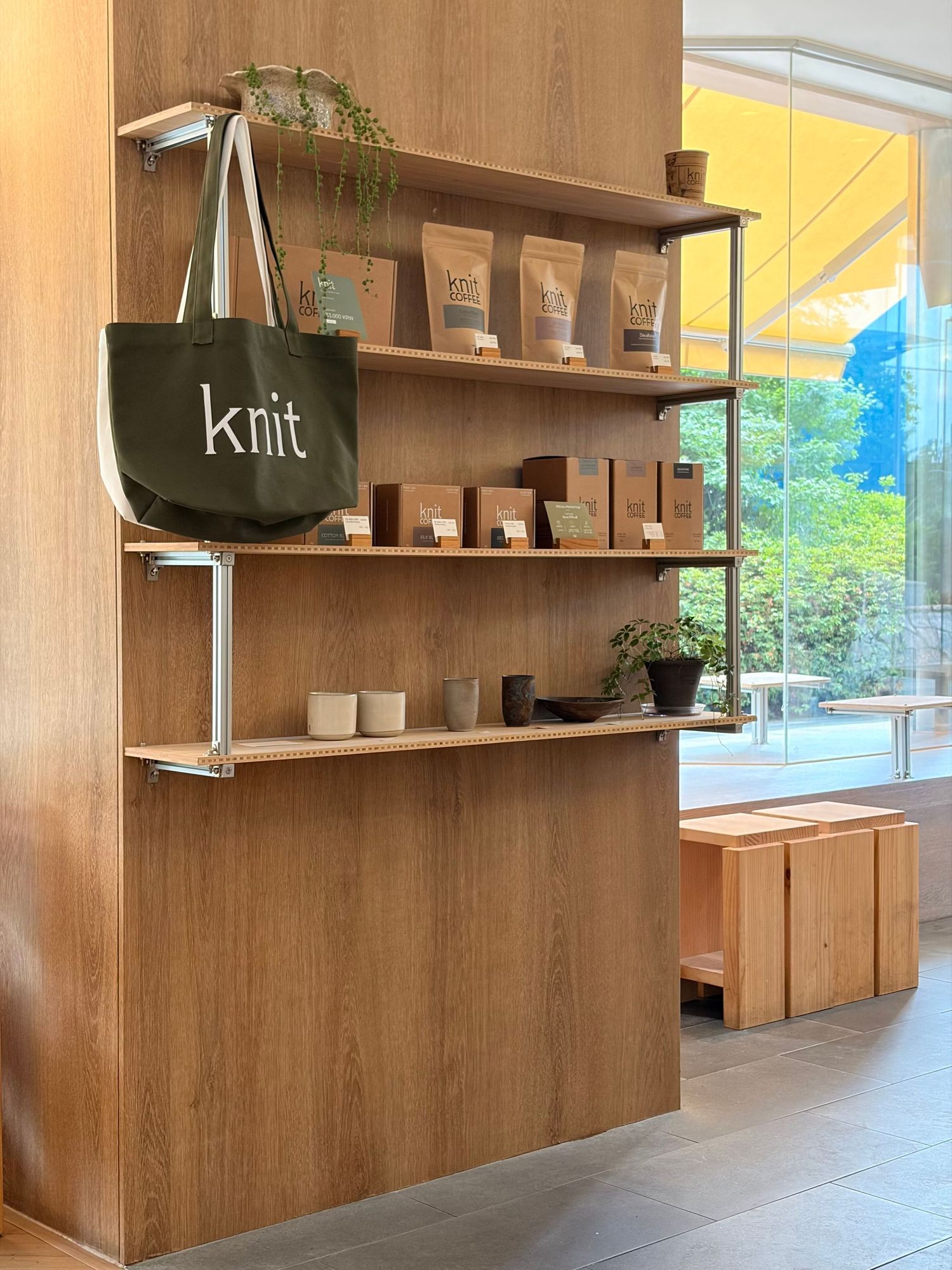 Wooden retail shelving display with Knit branded tote bag, coffee products, ceramic mugs, and small plants in a modern shop.