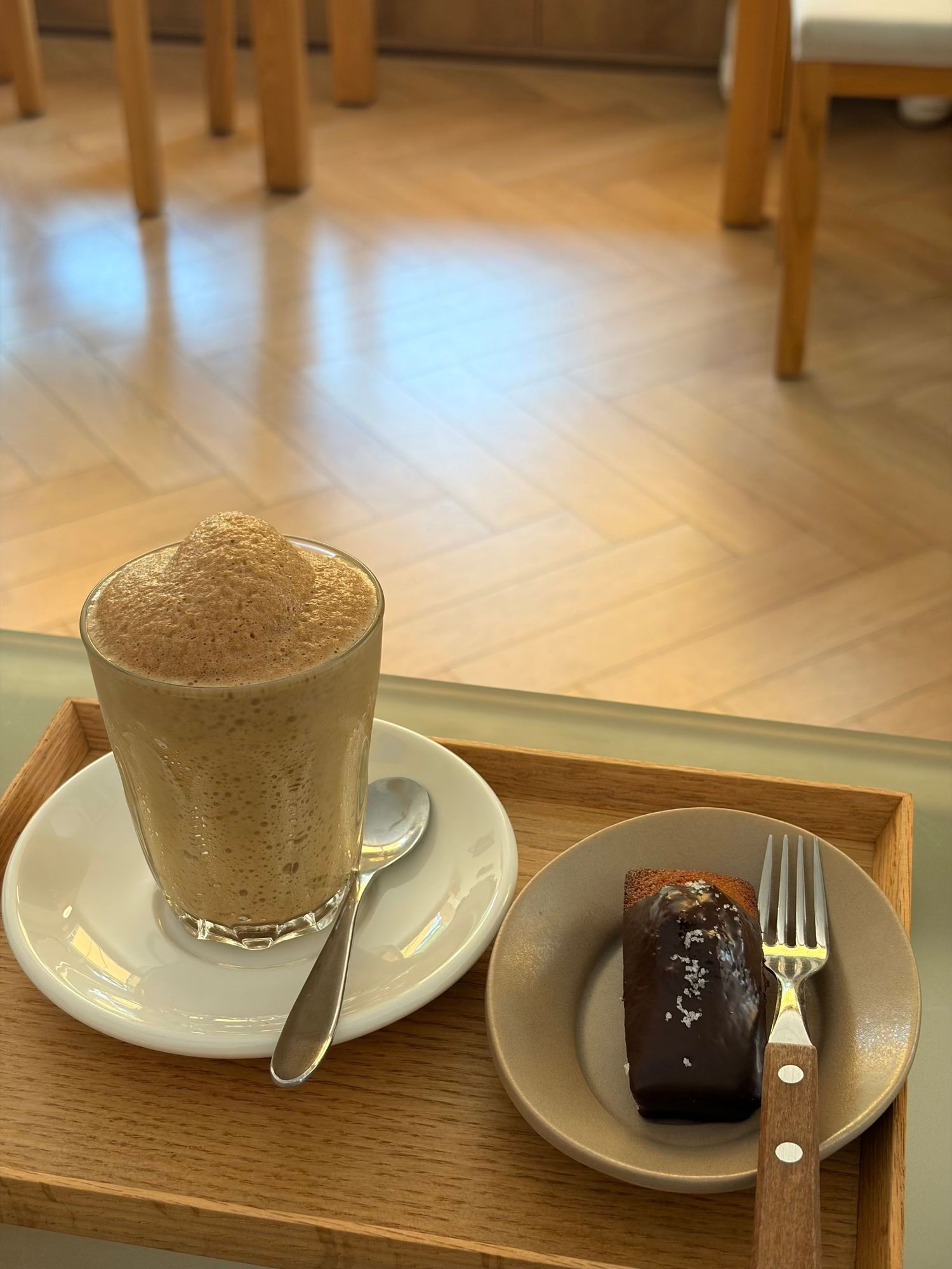 Frothy iced coffee in a tall glass beside a chocolate glazed pastry on a wooden tray in a cafe.