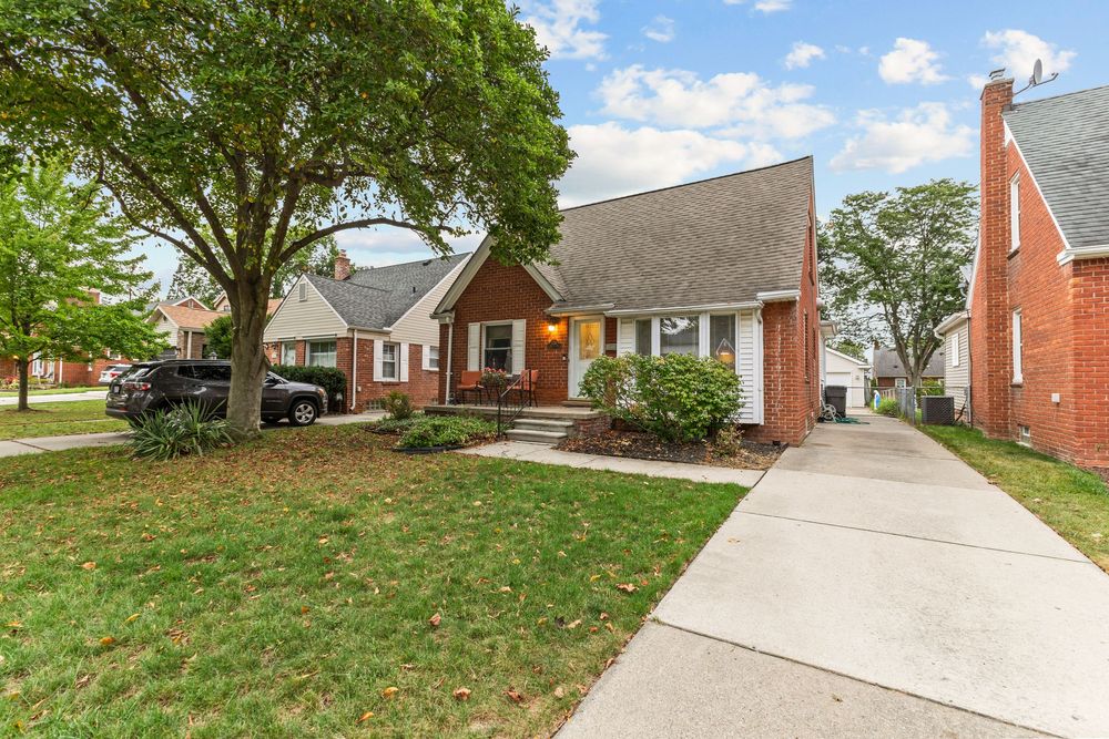 Charming brick Cape Cod home with mature trees, manicured lawn, concrete driveway, and classic architectural details.