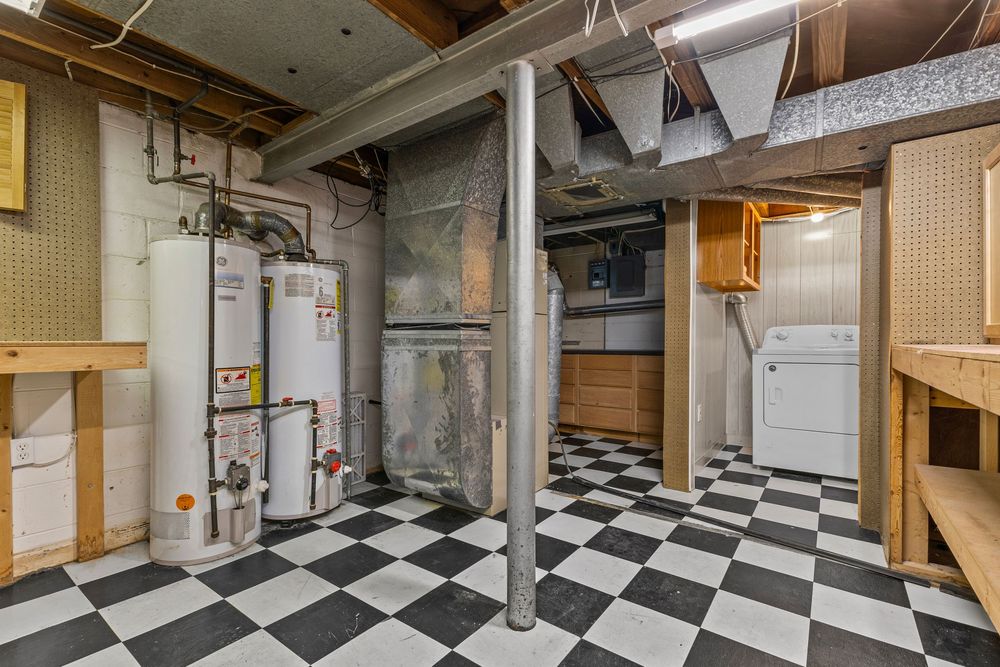 Unfinished basement with checkered black and white tile floors, water heater, support pole, and washer dryer unit.