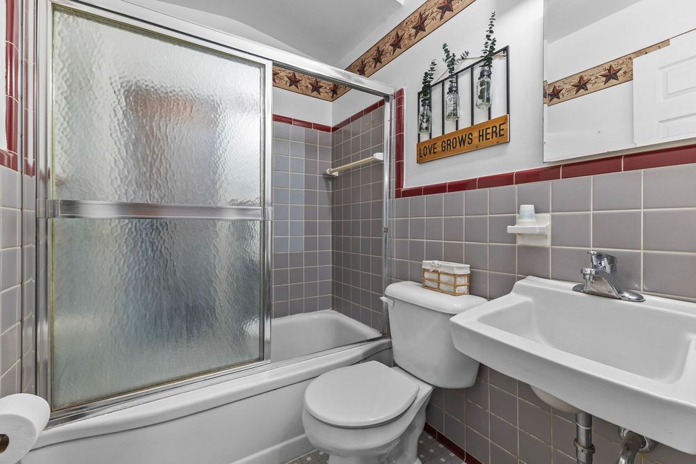 Compact bathroom featuring gray tile walls, glass sliding shower doors, pedestal sink, and decorative border tiles.