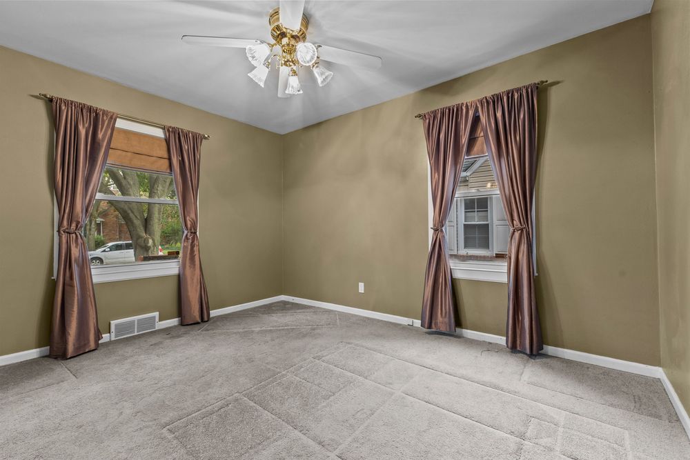 Spacious empty room with beige carpet, olive green walls, two windows with burgundy curtains, and a ceiling fan with light fixture.