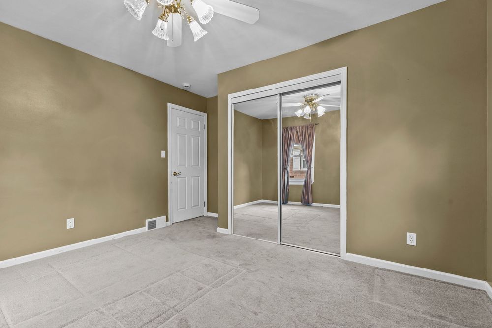 Carpeted bedroom with ceiling fan, mirrored closet doors, and neutral tan walls in a residential home.