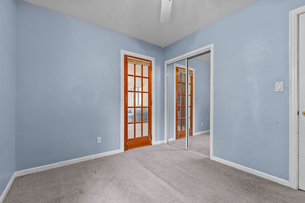 Light blue bedroom with gray carpet, wooden French doors, and mirrored closet doors in a bright, airy space.
