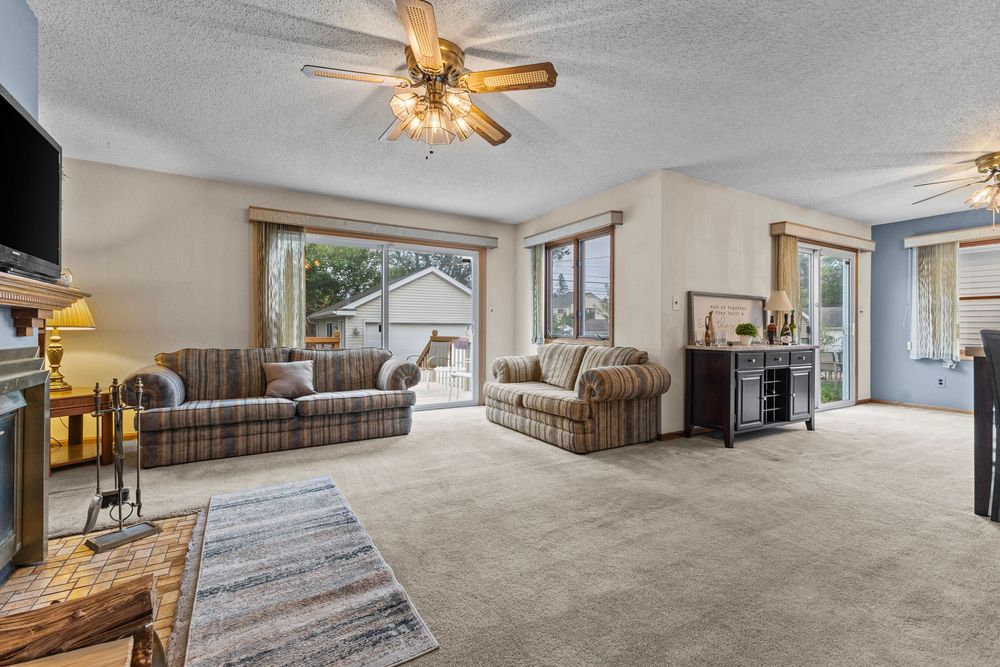 Spacious living room with plaid sofas, ceiling fan, large windows, and neutral carpet flooring.