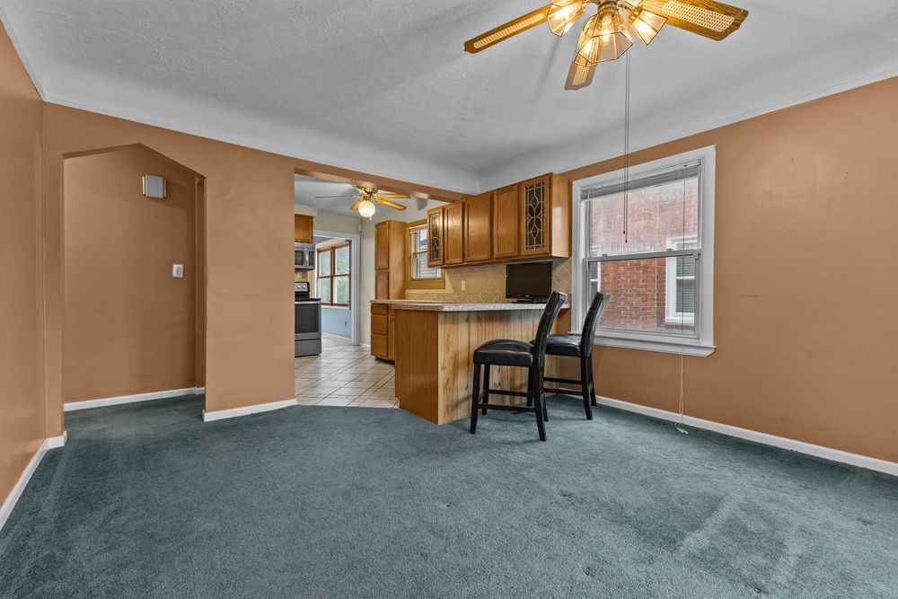 Open dining area with blue carpet, bar seating, kitchen peninsula, ceiling fan, and natural light from a window.