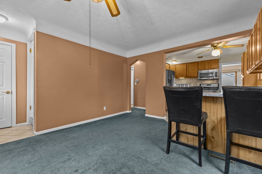 Dining area with teal carpet, tan walls, ceiling fan, and view into kitchen with bar seating and stainless appliances.