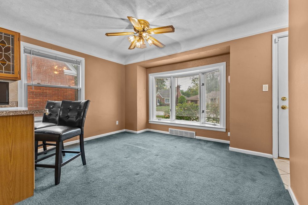 Bright dining area with teal carpet, ceiling fan, large windows, and a black bar-height chair near a kitchen counter.