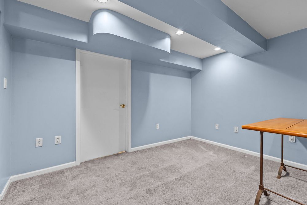 Bright basement room with light blue walls, gray carpet, recessed lighting, and a wooden drafting table in the corner.