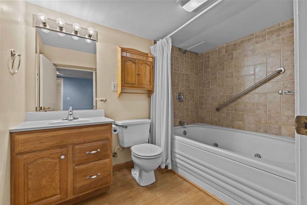 Bright bathroom with wood vanity, large mirror, tiled shower-tub combo with curtain, toilet, and light wood floors.