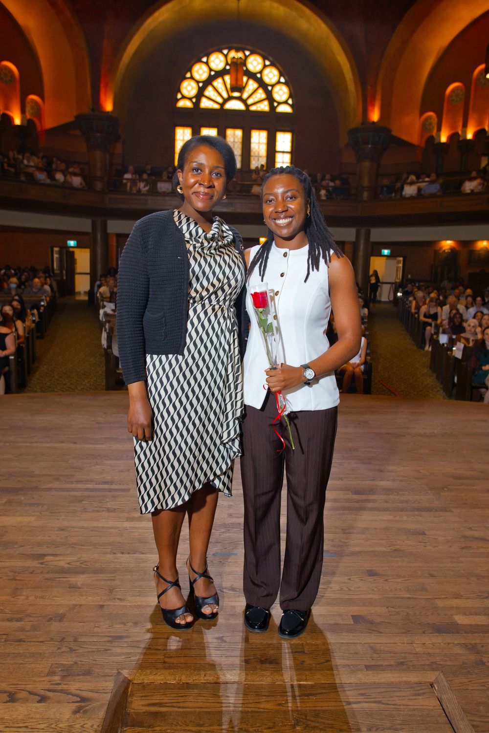 Two women pose together smiling at a formal ceremony in an ornate hall, one holding a red rose.