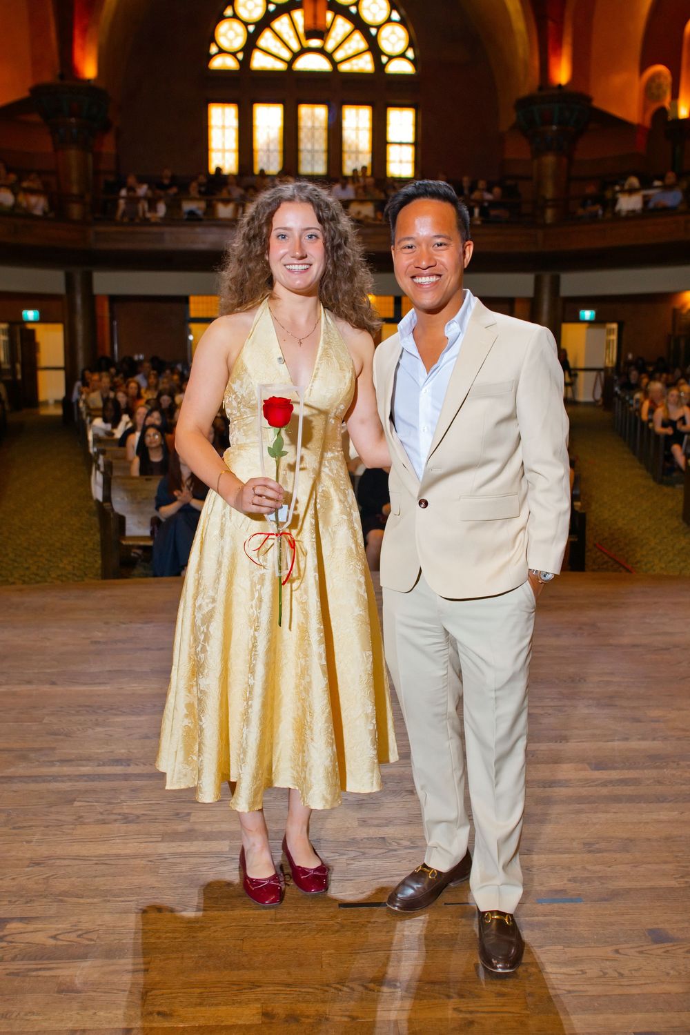 A woman in a gold dress holding a red rose poses with a man in a beige suit in an elegant hall.