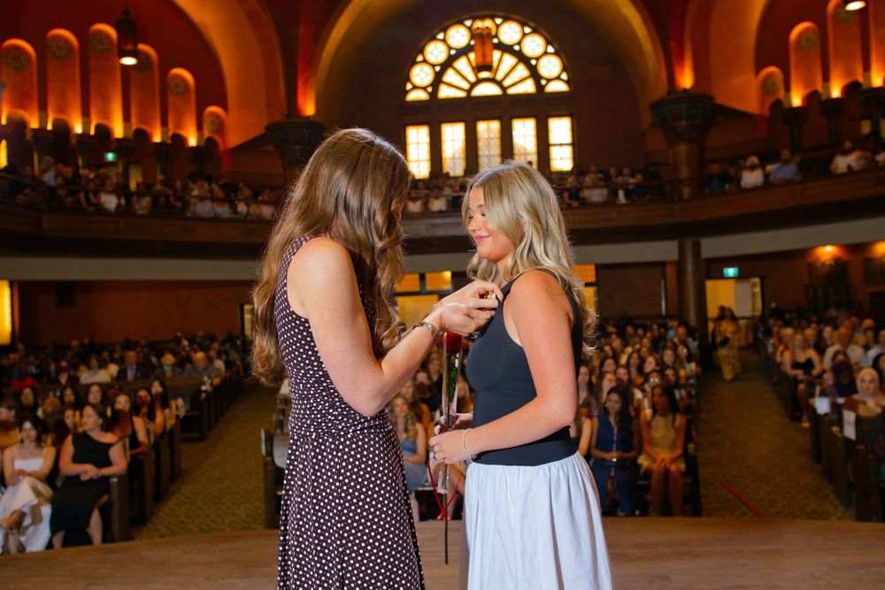 Two women share a meaningful moment on stage during a formal ceremony in an ornate auditorium filled with attendees.