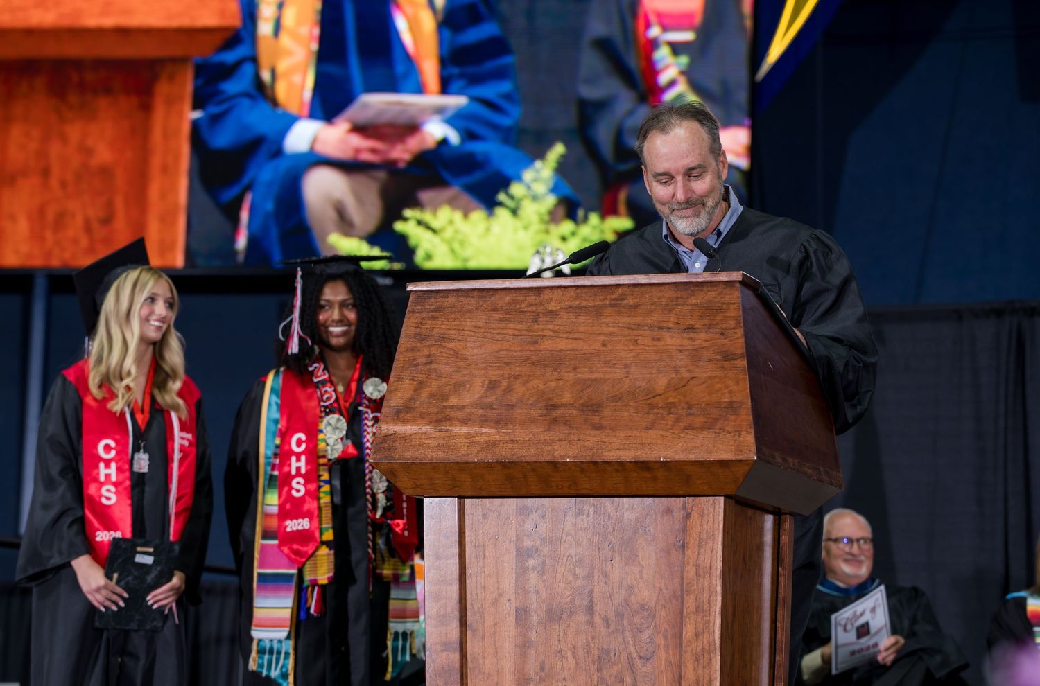 Man speaking at wooden podium during graduation ceremony with students in red sashes standing nearby.