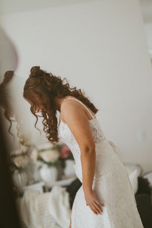Bride in lace wedding dress with curly hair bending forward during bridal preparation in a softly lit room.