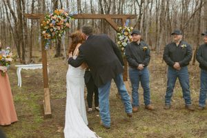 Outdoor woodland wedding ceremony with bride and groom embracing under a floral arch, guests in jeans watching nearby.