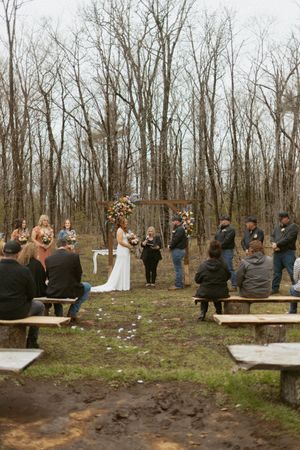 Outdoor woodland wedding ceremony with bride and groom at floral arch, guests seated on rustic benches among bare trees.