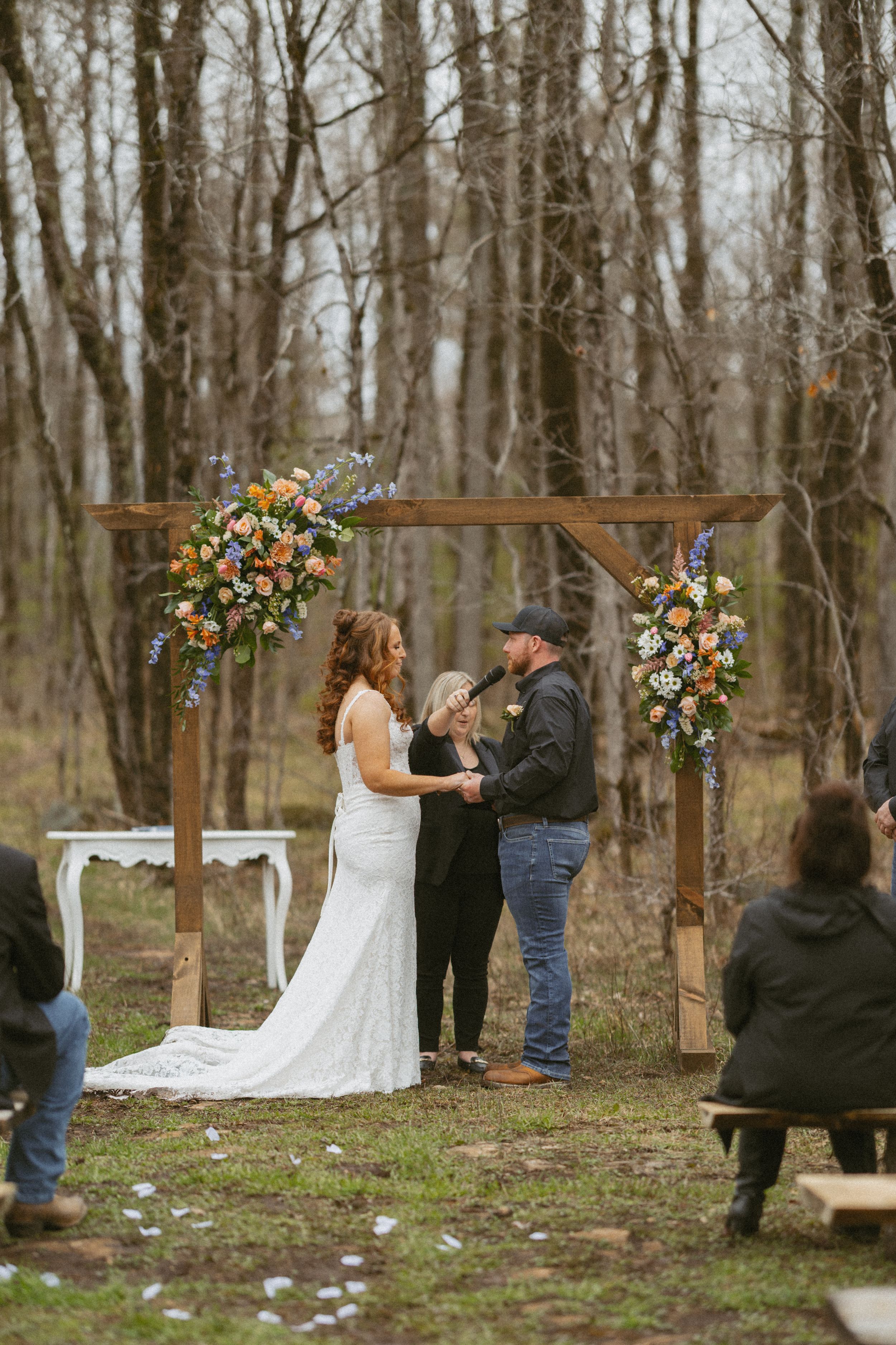 Bride and groom exchange vows under a rustic wooden arch adorned with colorful flowers in a woodland wedding ceremony.