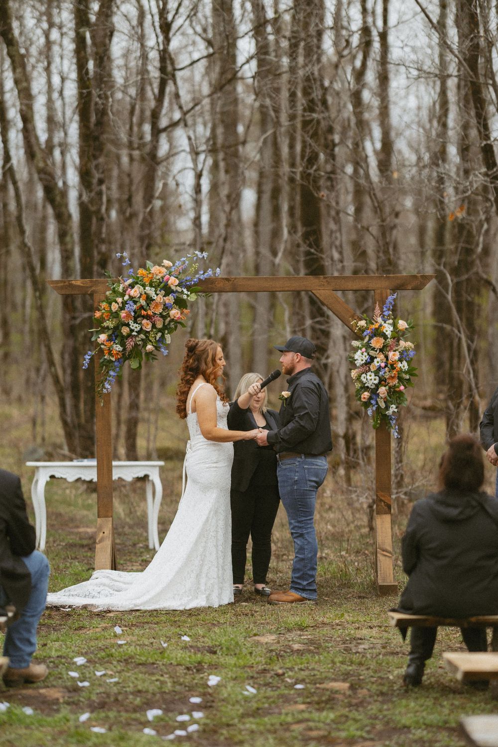 Bride and groom exchange vows under a rustic wooden arch adorned with colorful flowers in a woodland wedding ceremony.