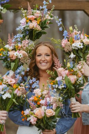 Smiling bride framed by colorful wedding bouquets featuring pink, blue, and peach flowers held by bridesmaids.