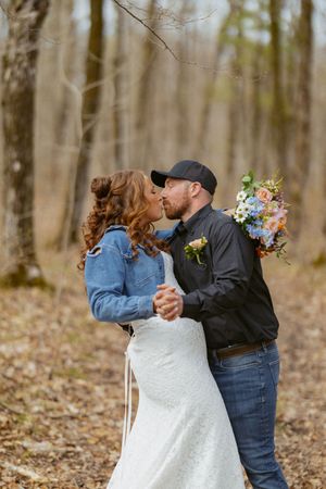 Newlywed couple shares a kiss in a forest setting, bride in white lace gown holding a pastel bouquet.