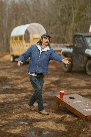 Man in denim jacket playing cornhole on muddy ground near a covered wagon and truck in a wooded area.