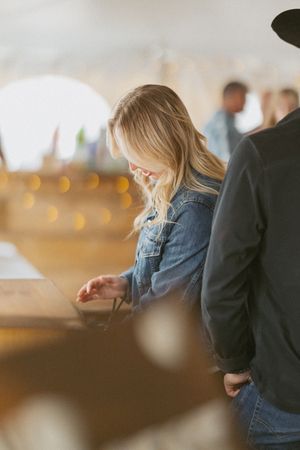 Blonde woman in denim jacket looks down at a wooden table inside a warmly lit wedding reception tent.