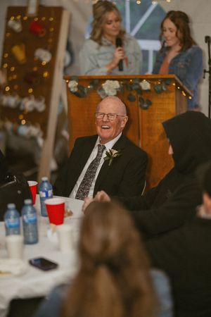 Elderly man in suit and tie laughing joyfully at a wedding reception while guests and speakers surround him.