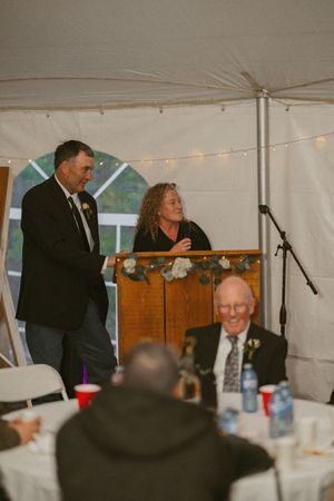 Couple speaking at a wedding reception podium inside a tent with string lights and floral decorations.