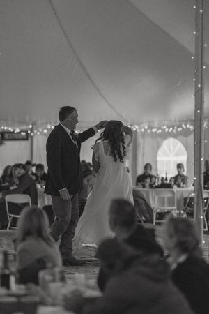 Black and white photo of a bride and groom sharing their first dance under a tent adorned with string lights.