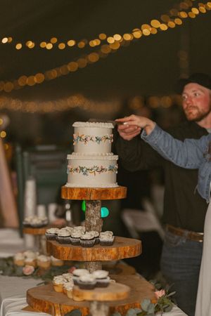 Man placing topper on rustic three-tier wedding cake on wooden stand with cupcakes, under warm string lights.