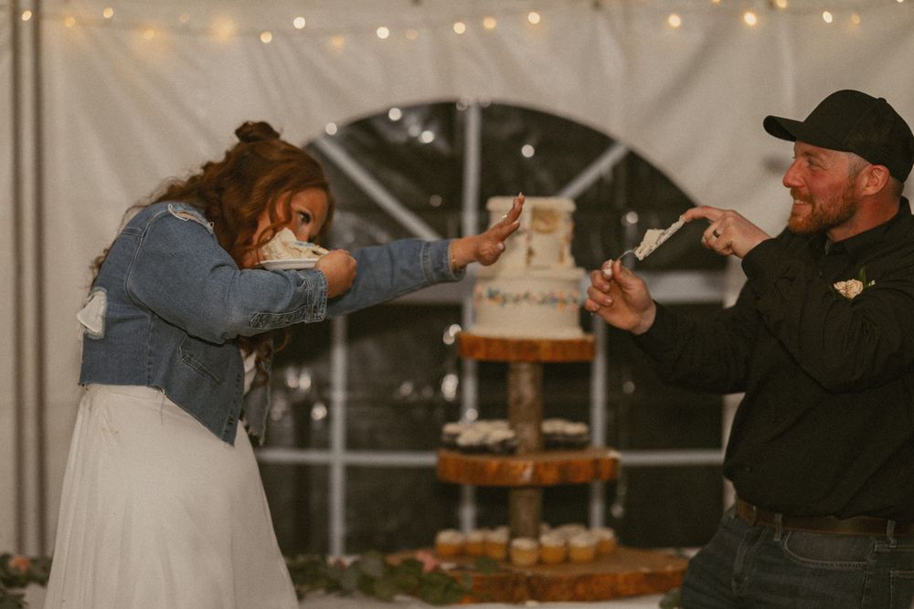 Bride and groom playfully feeding each other wedding cake at rustic tented reception with fairy lights.