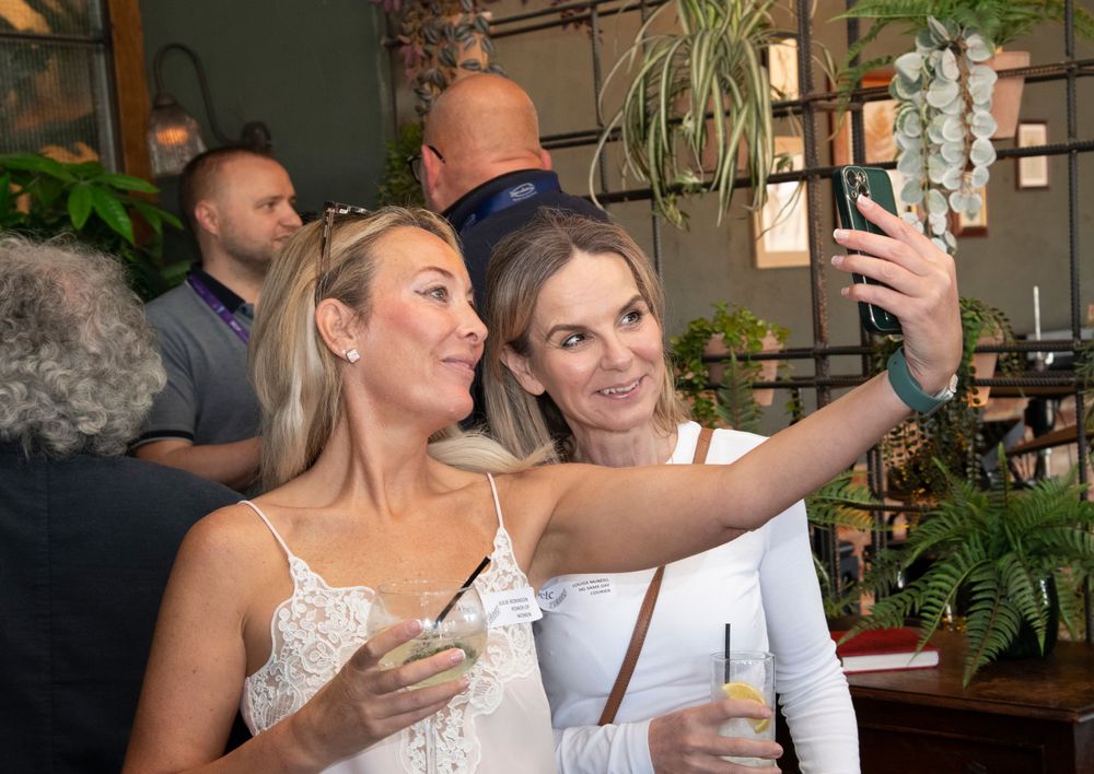Two women in white outfits taking a selfie with cocktails at a social gathering in a plant-filled venue.