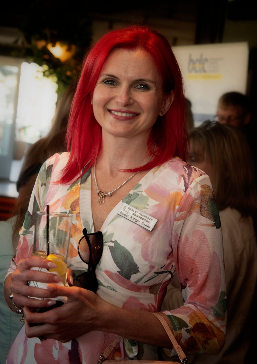 Woman with vibrant red hair smiling at event, holding a drink and sunglasses, wearing a floral dress and name tag.