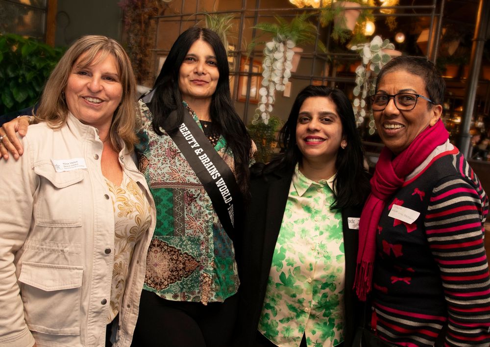 Four women posing together at an indoor event, one wearing a pageant sash, smiling for a group photo.