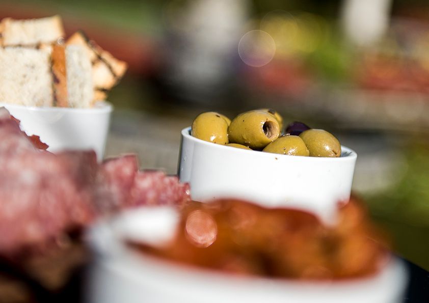 Close-up of green olives in white serving bowl with blurred appetizers in background.