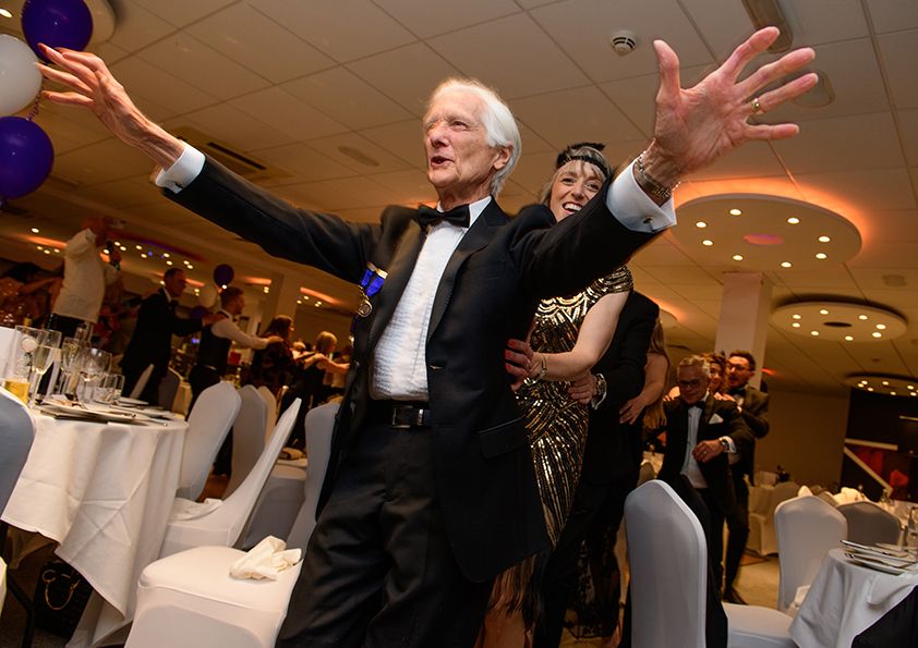 A person in a tuxedo gestures dramatically during a formal event with purple balloons.