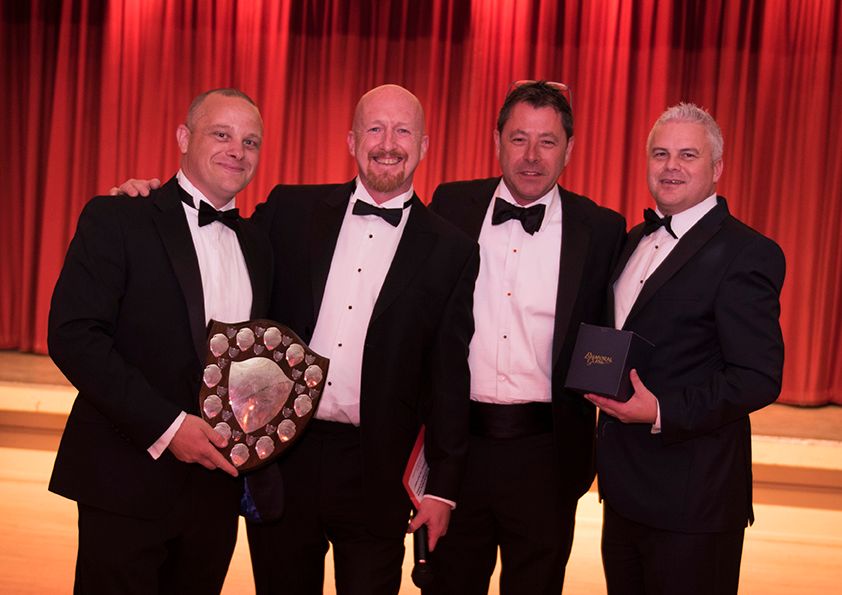 Four men in formal black tie attire pose with awards at an elegant event with red curtain backdrop.