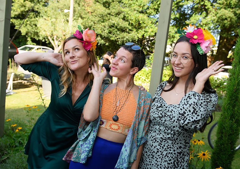 Three women in colorful dresses pose playfully with floral headpieces at an outdoor event.