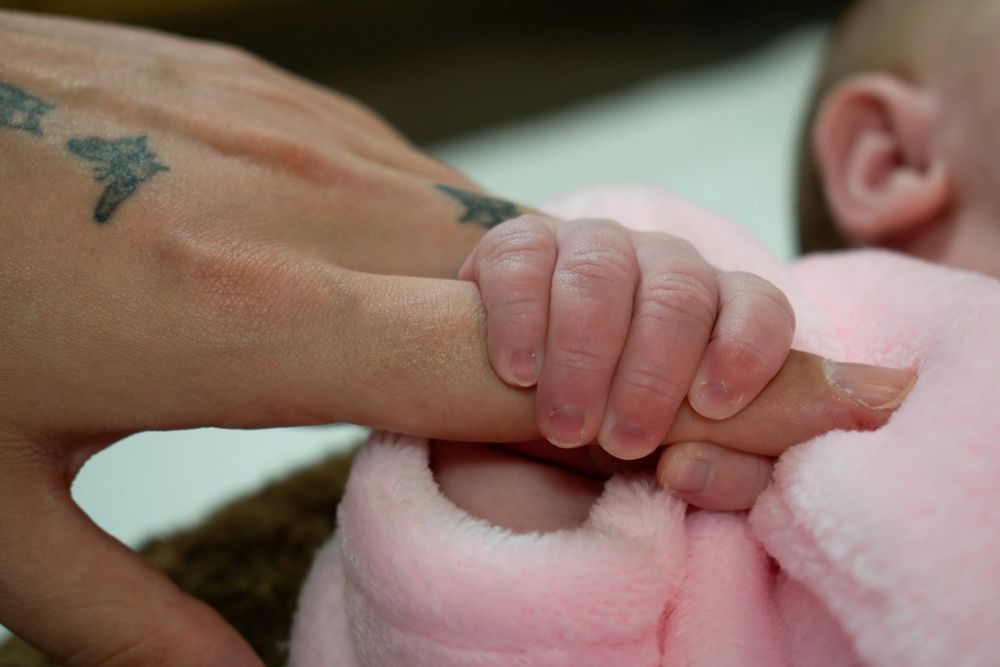 A tattooed adult hand gently holding a tiny newborn baby's hand on a pink blanket.