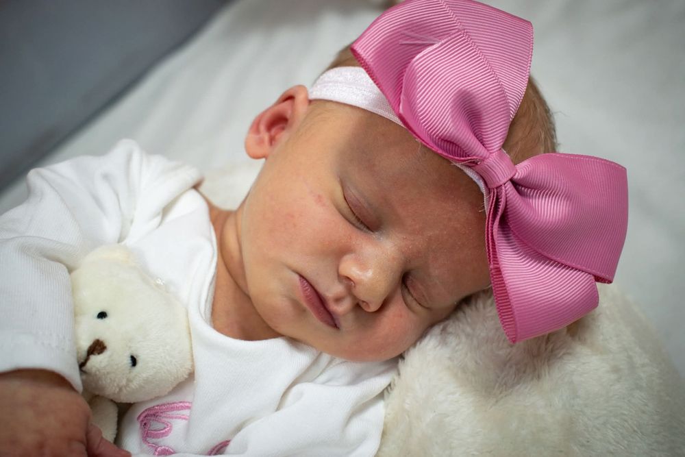 Sleeping newborn baby girl with a large pink bow headband cuddling a small teddy bear.