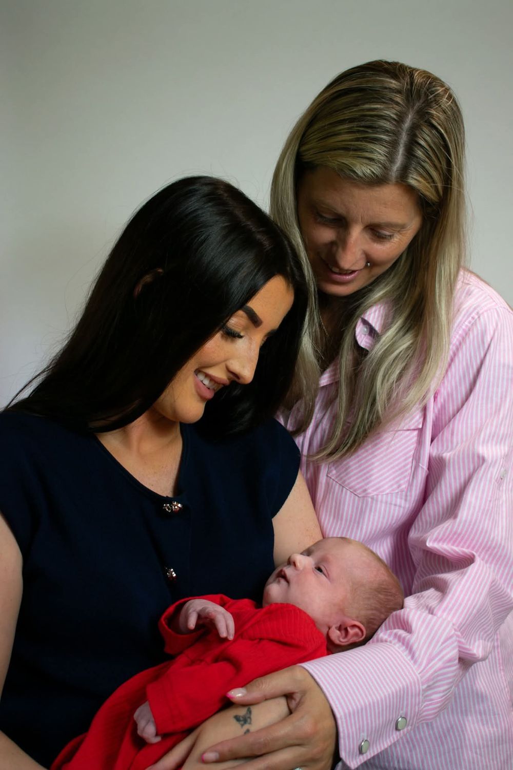 Two women smiling and bonding over a newborn baby wrapped in a red blanket.