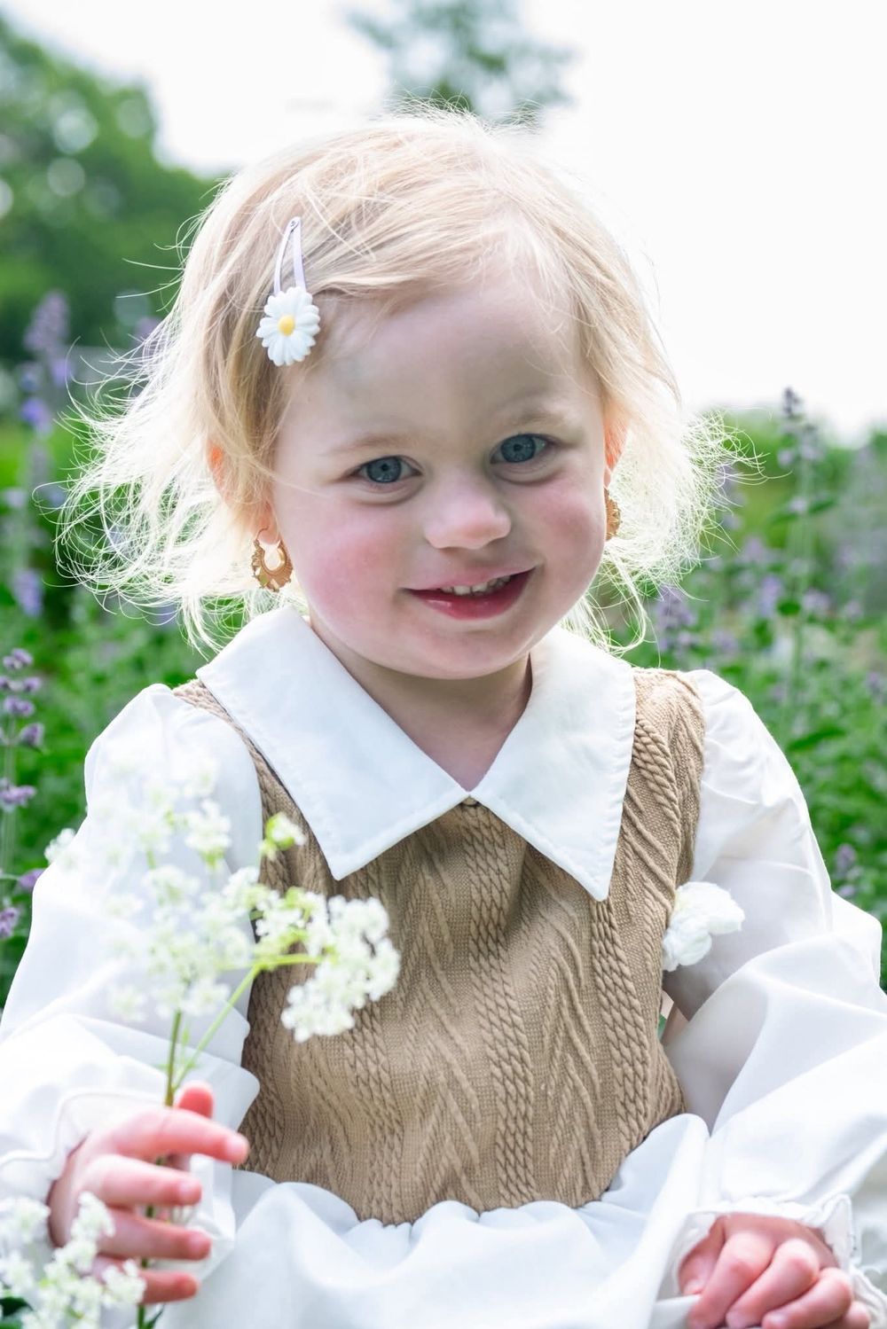 Adorable toddler girl with flower hair clip holding white wildflowers in a garden.